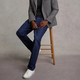 Person wearing a grey blazer, black shirt, blue jeans, and white training shoes on a wooden stool against a grey background.